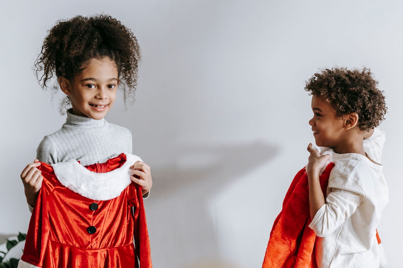 Cheerful African American siblings with red Santa Claus outfits in hands standing in light room at home during Christmas holiday